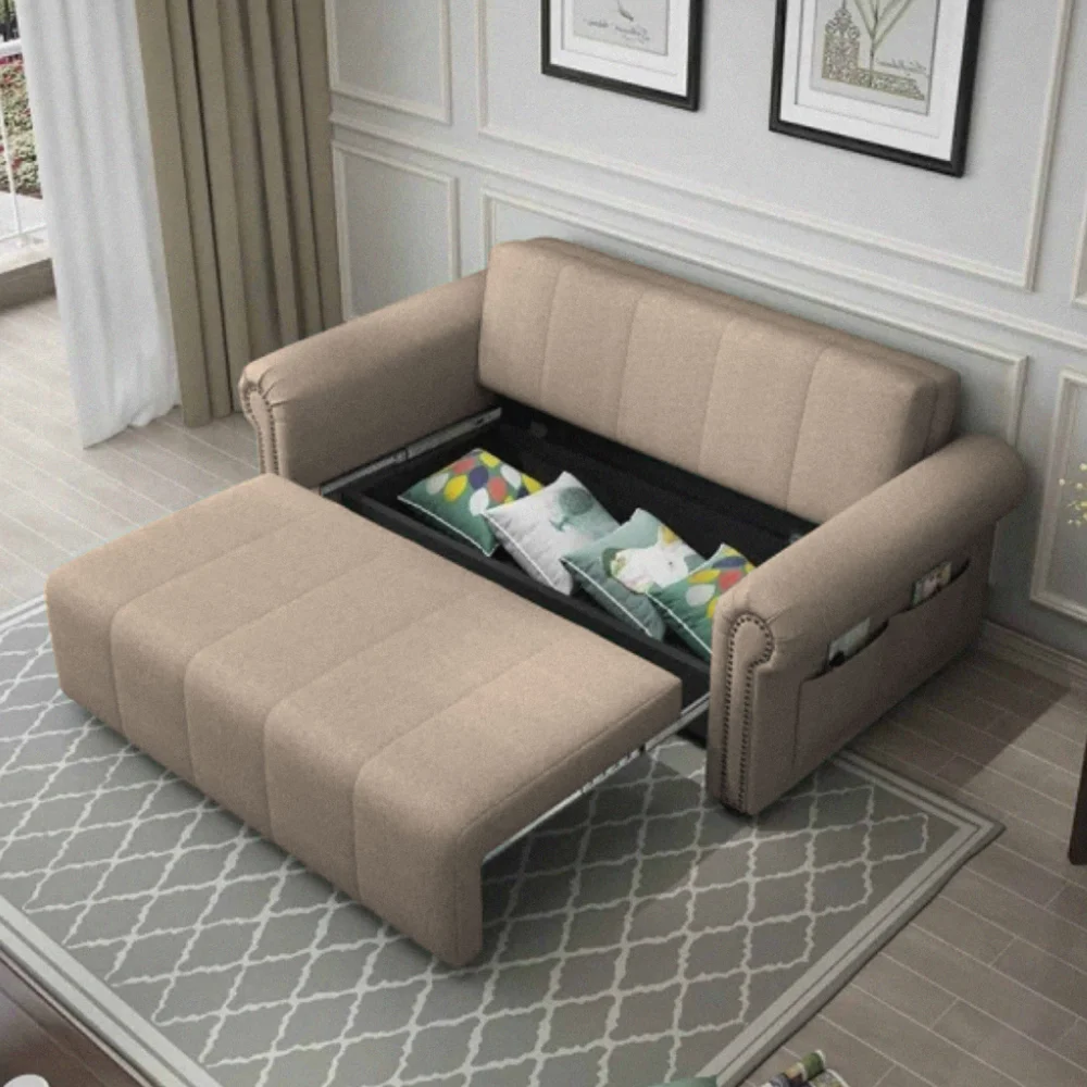 A light beige pull-out sofa bed in a classic living room setting, extended to show the sleeping surface and internal storage for decorative pillows, featuring armrest storage pockets
